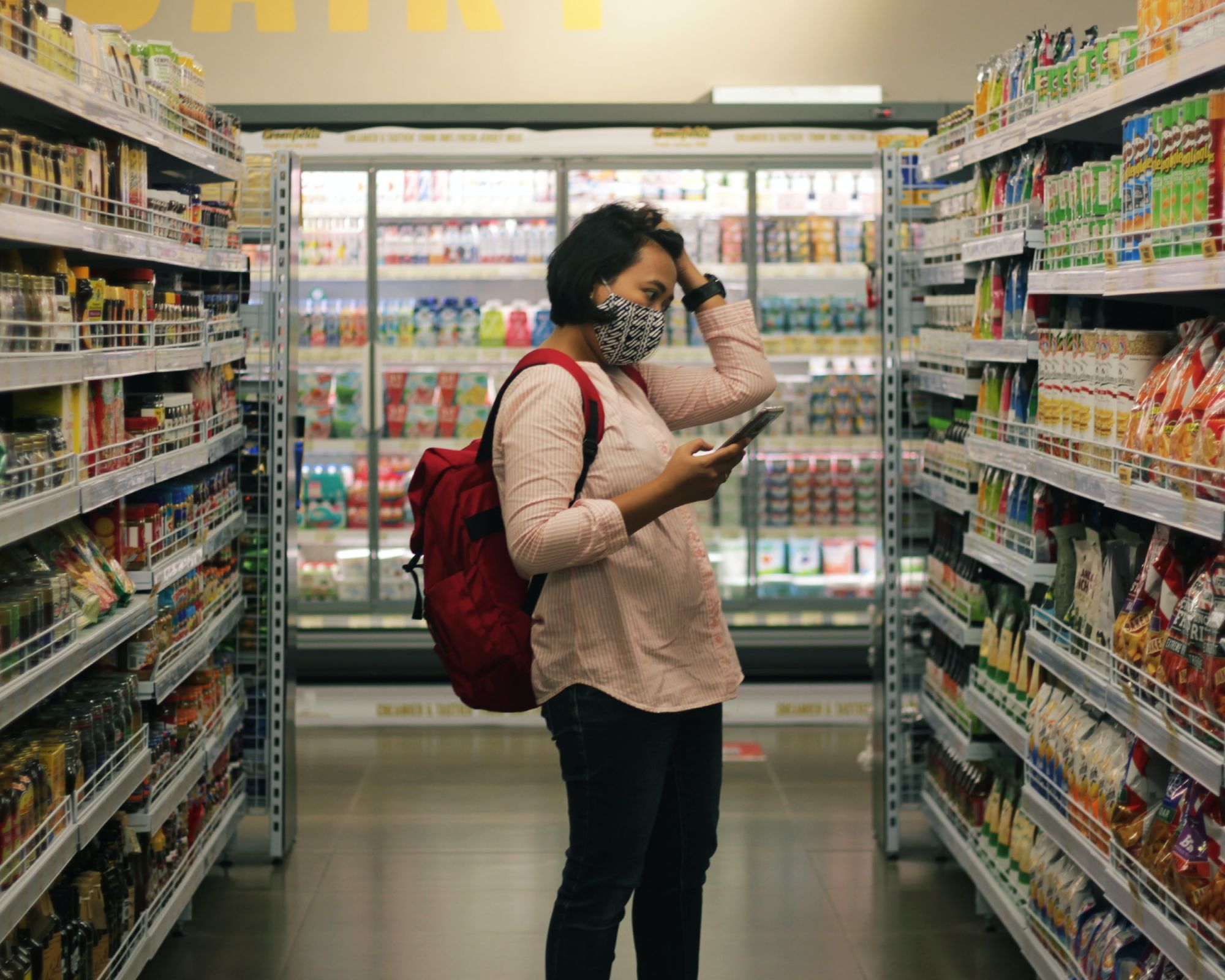 Frau im Supermarkt mit vollen Regalen an den Seiten und Kühlschränke im Hintergrund